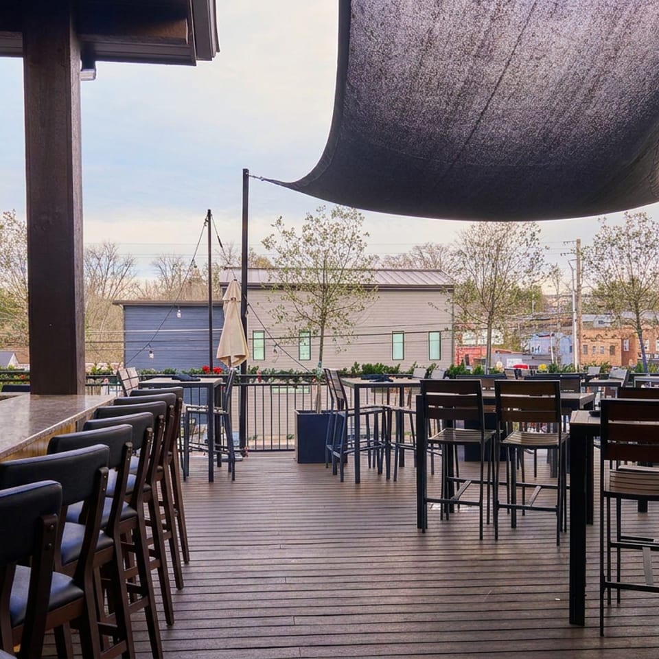 Spacious rooftop restaurant patio with bar seating, tables, shade sails, string lights, and views of surrounding buildings and trees.