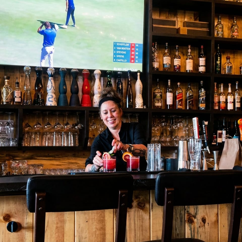 Bartender prepares two red cocktails behind a wooden bar lined with liquor bottles, glassware, and sports television.