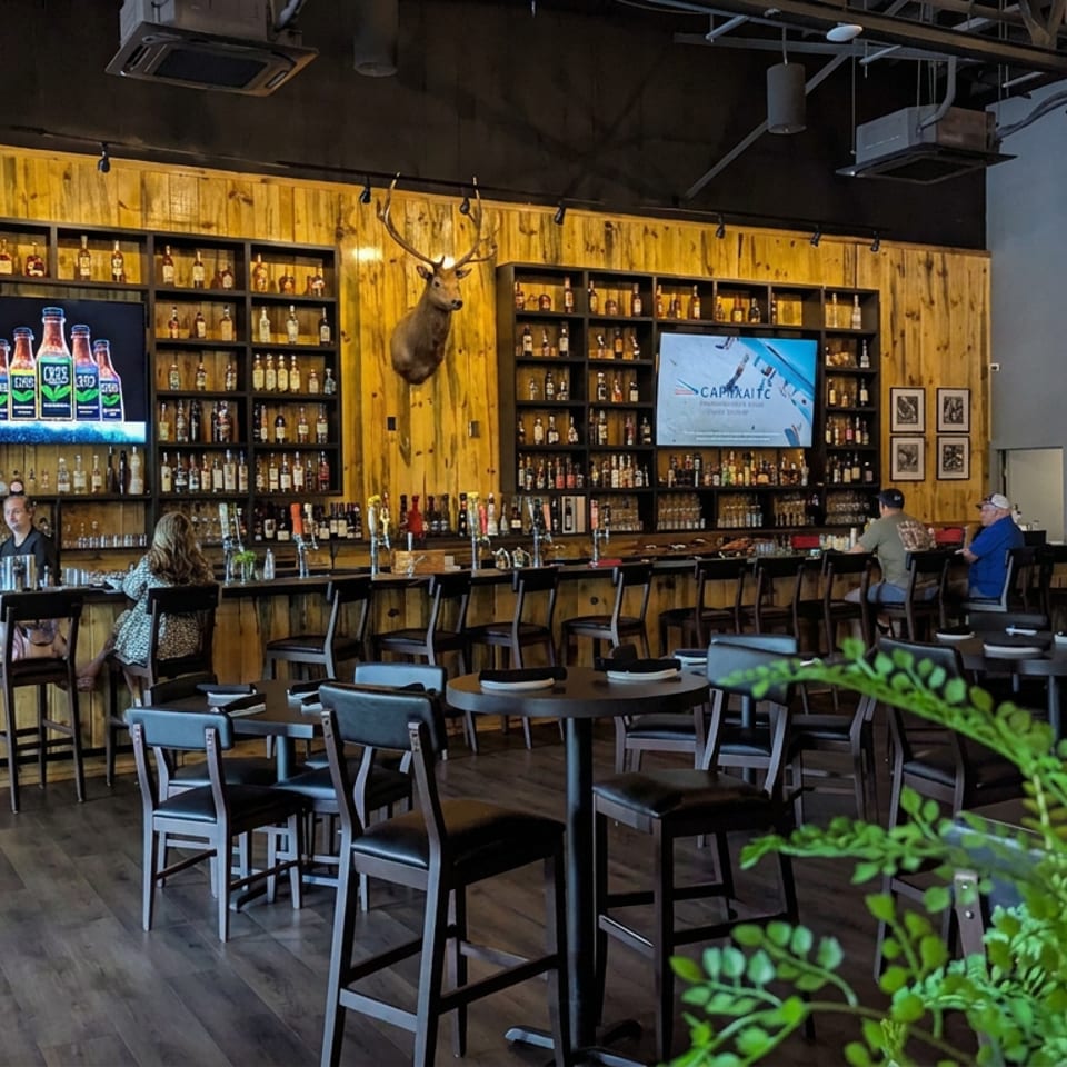 Rustic restaurant bar with wood-paneled walls, liquor shelves, mounted deer head, televisions, and patrons seated at tables.