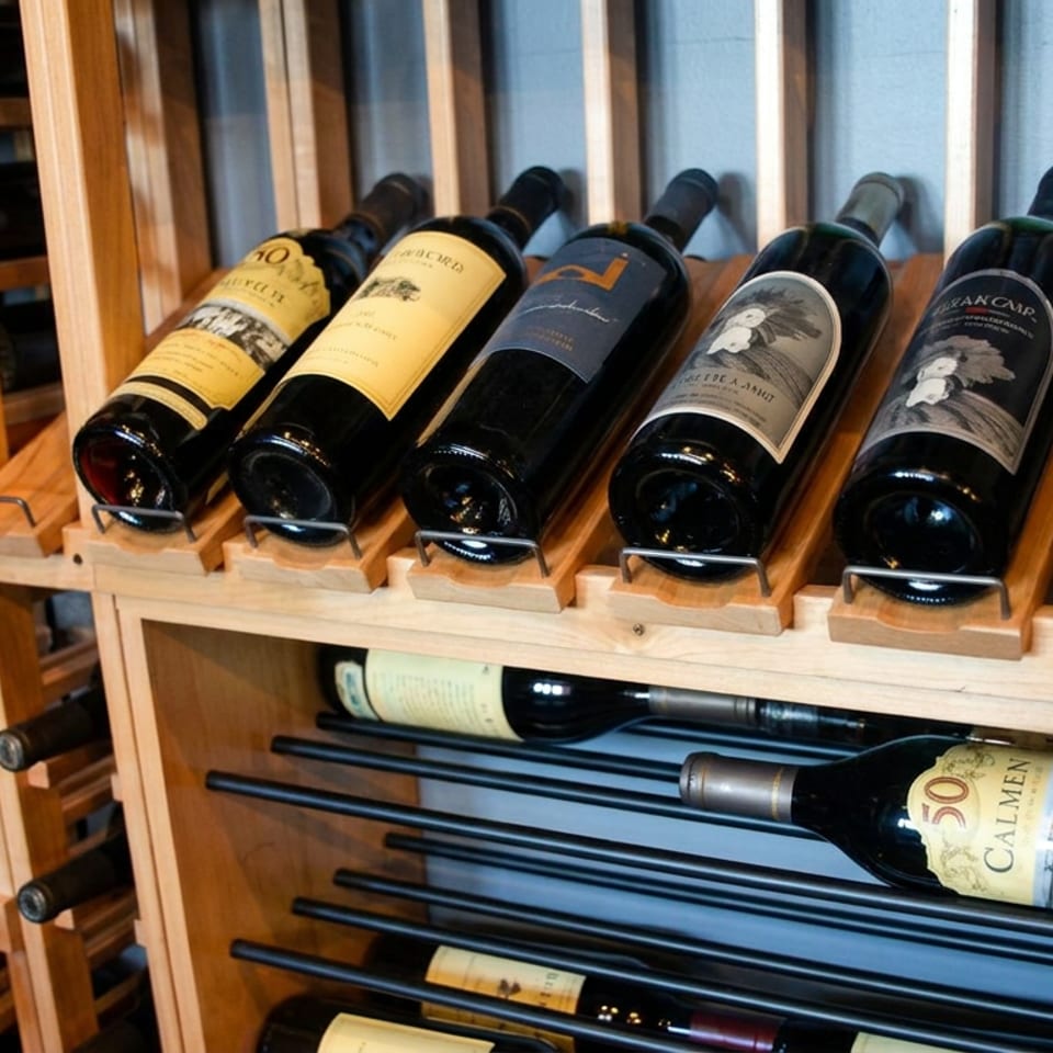 Wooden wine rack displaying rows of horizontally stored wine bottles, including a prominent Carmenère bottle.