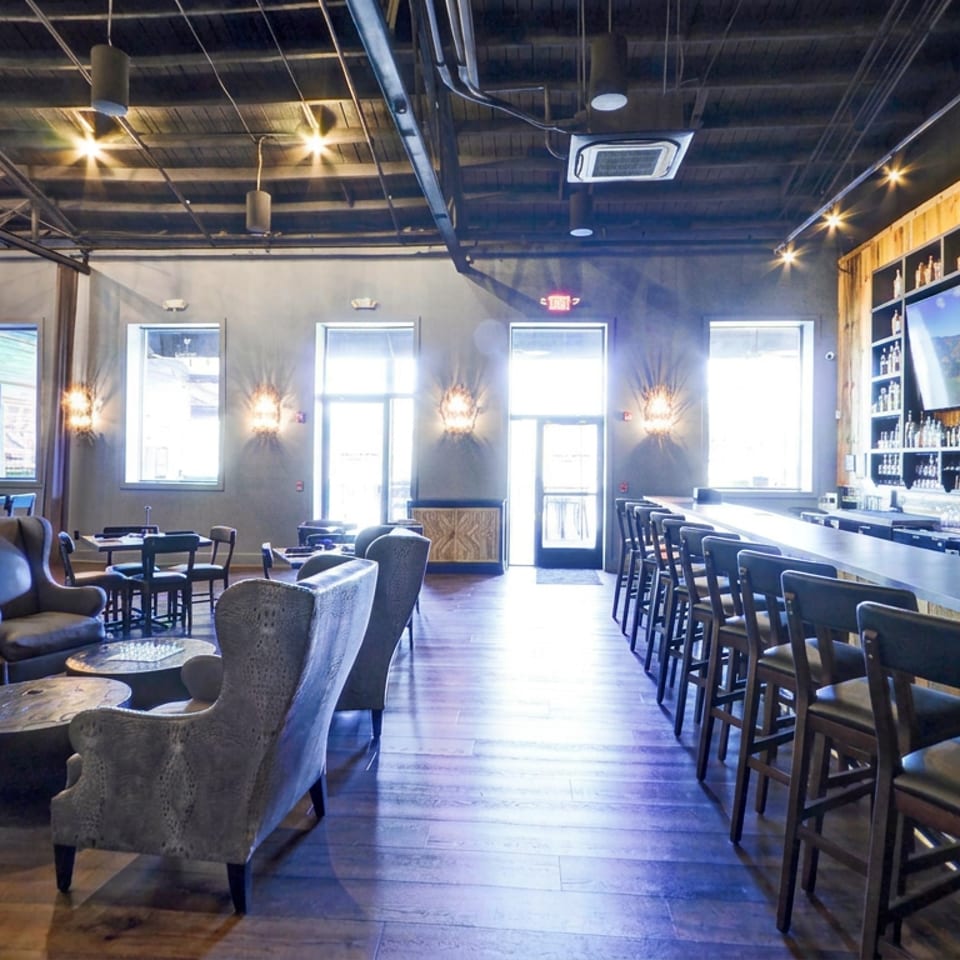Spacious modern restaurant bar with extensive liquor shelves, televisions, counter seating, lounge chairs, and exposed industrial ceiling.