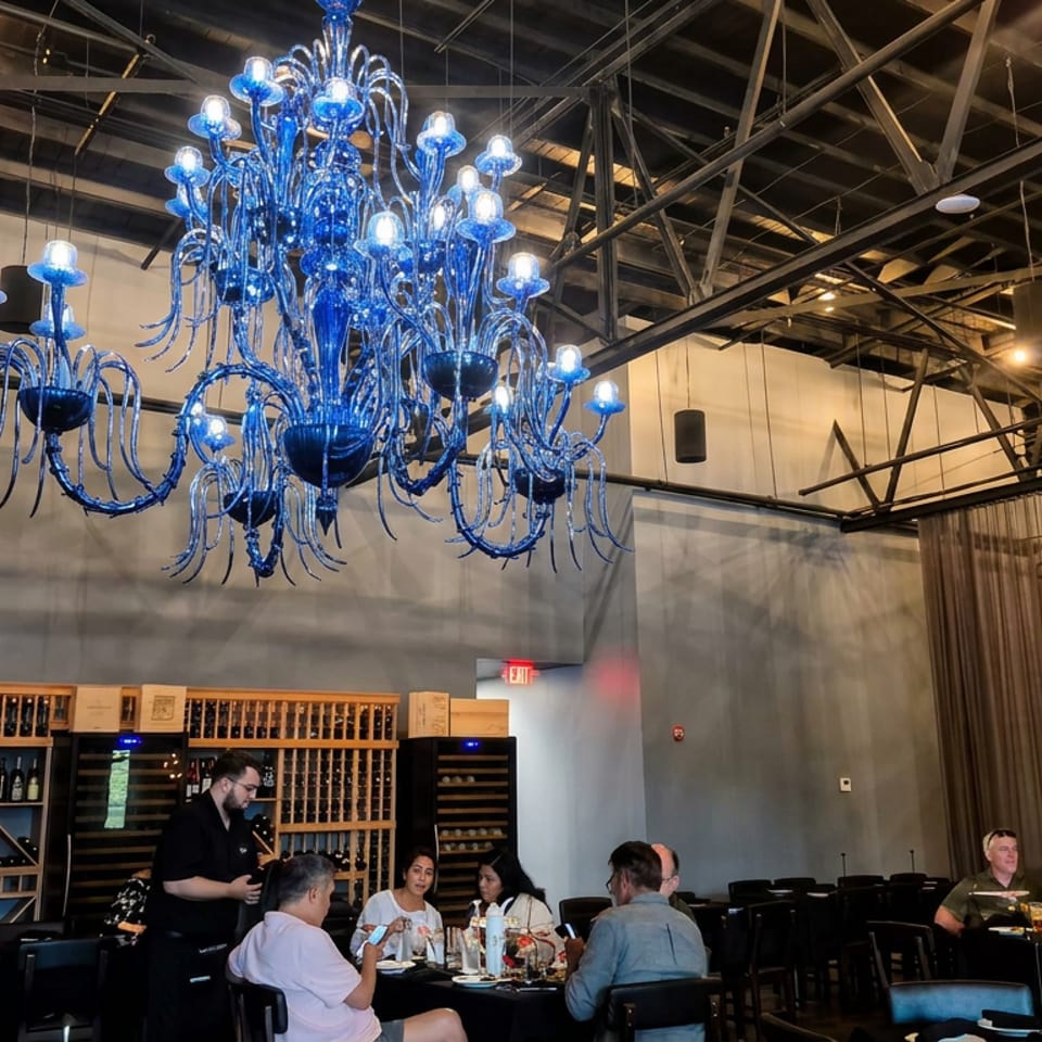 Industrial-style restaurant dining room featuring a dramatic blue glass chandelier, wine racks, and seated diners.