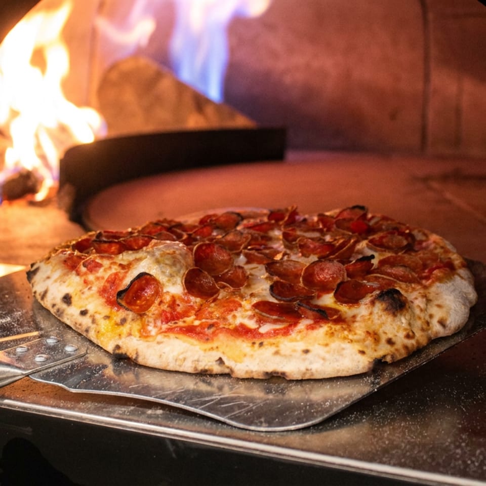 Pepperoni pizza on a metal peel baking inside a wood-fired oven with flames glowing behind it.