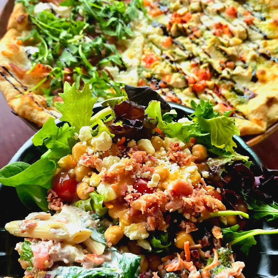 Flatbread pizza topped with greens and vegetables alongside a colorful mixed salad on black plates.