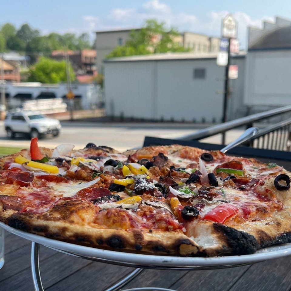 Loaded pizza topped with pepperoni, sausage, olives, peppers, onions, and cheese on an outdoor restaurant table overlooking a street.